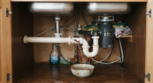 Close up photograph of a damaged PVC water pipe leaking under a clean stainless steel sink inside an apartment kitchen, realistic documentary lighting in soft tones